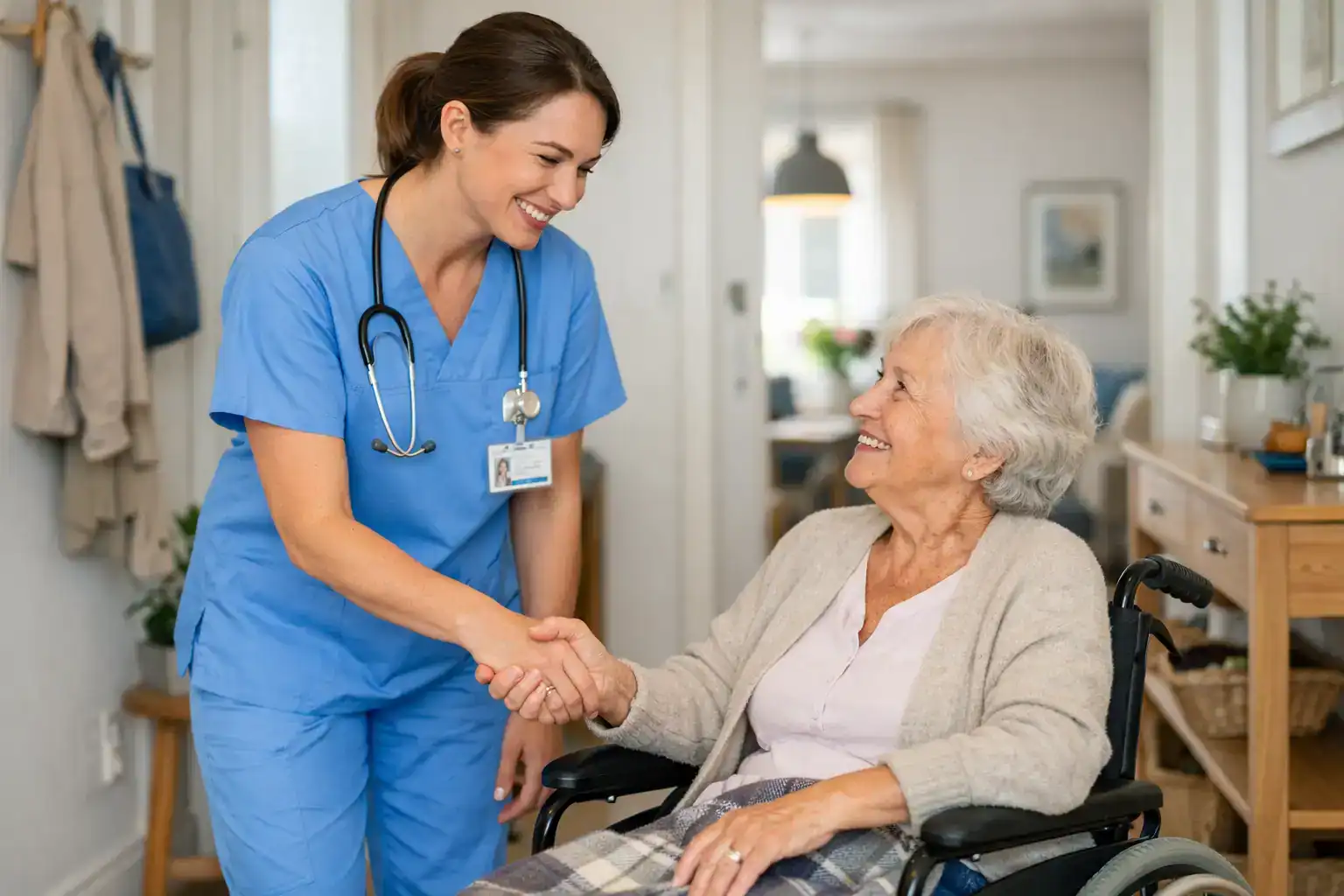 Pflegefachkraft in blauer Arbeitskleidung begrüßt eine ältere Dame im Rollstuhl mit Handschlag in einer häuslichen Umgebung.