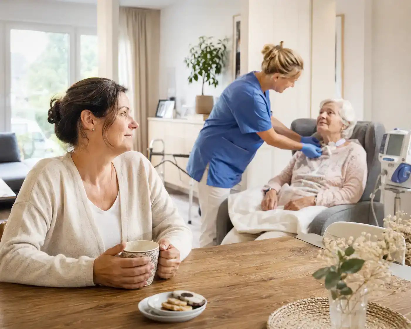 Angehörige sitzt mit einer Tasse am Esstisch, während eine Pflegefachkraft im Hintergrund eine Patientin mit Tracheostoma zu Hause versorgt.