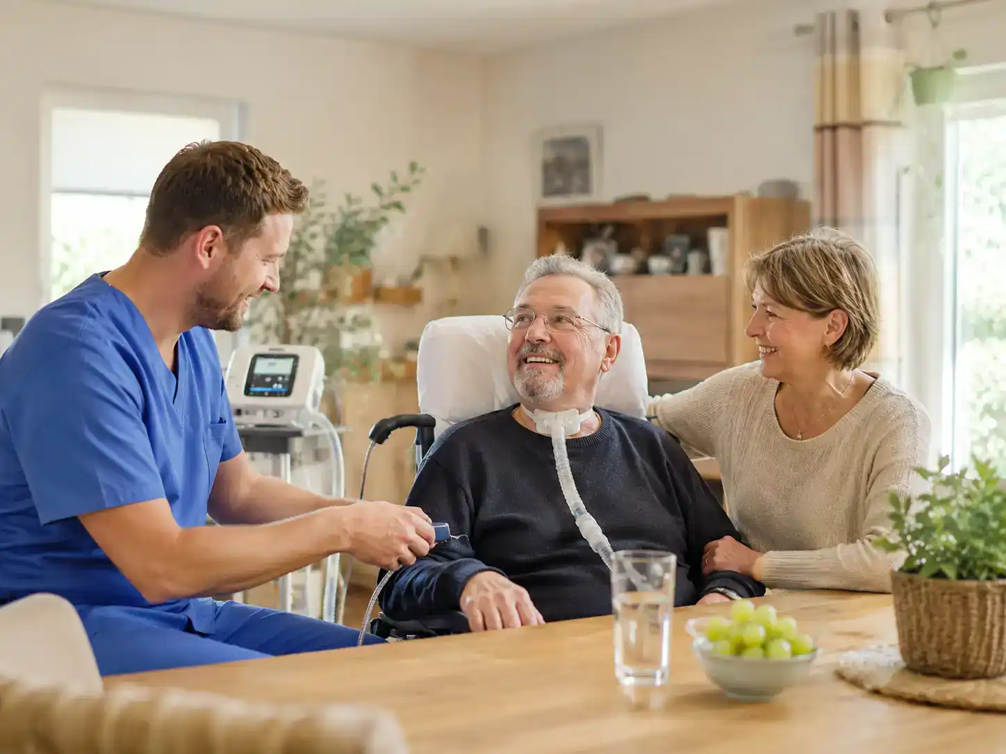 Pflegefachkraft betreut einen Patienten mit intensivpflegerischem Bedarf und spricht mit ihm und einer Angehörigen in einem wohnlichen Umfeld.